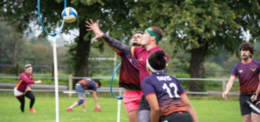 There is a keeper in a red jersey and a chaser in a purple jersey. Both are reaching for the ball, which is already in the air in front of the hoop. Also visible is another chaser and two beaters in purple jerseys and one beater in a red jersey.