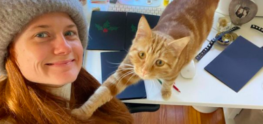 Bonnie Wright is smiling at the camera and her cat is reaching out to her. There are homemade Christmas cards on the desk.