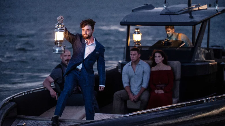 In a still from "The Lost City" from Paramount Pictures, Daniel Radcliffe can be seen in character as Fairfax. He is holding a lantern at the bow of a boat as Sandra Bullock and Channing Tatum's characters, seated on the boat behind him, look on.