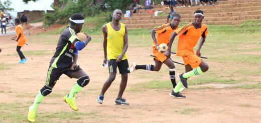 There is one chaser holding a ball and running forward, while other chaser and beater from opposite team is running next to him The beater is holding football ball (probably bludger). There is a refferee standing there also.