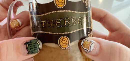 A hand with a Butterbeer manicure holds a Butterbeer glass