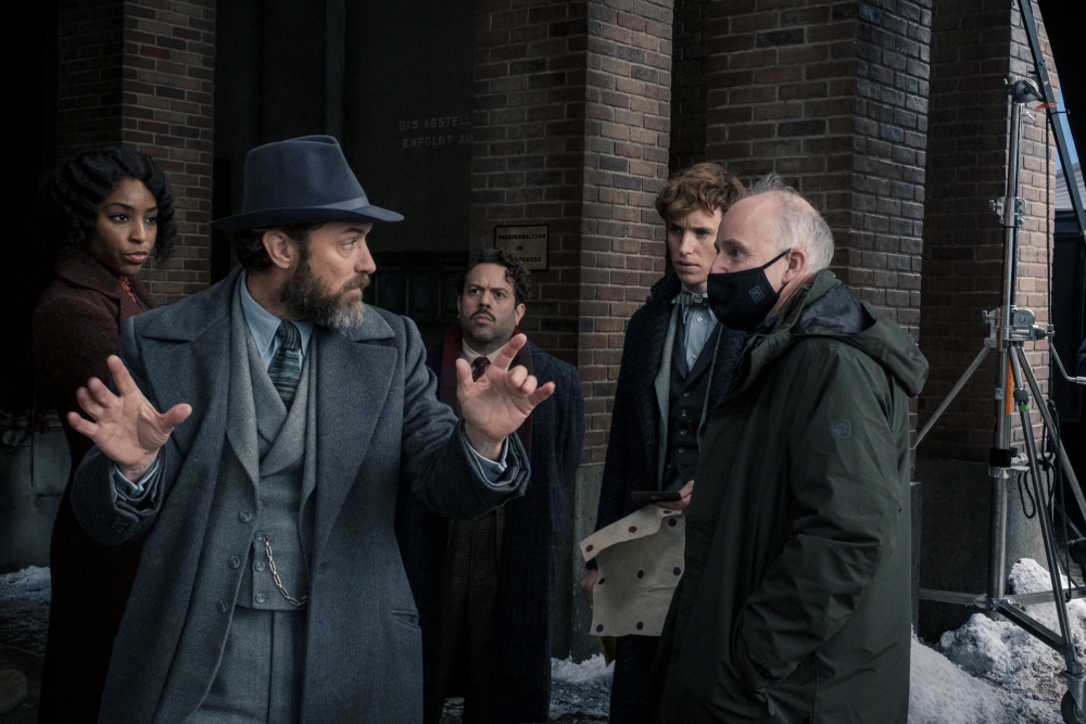 Director David Yates is shown conversing with actor Jude Law (Albus Dumbledore) on the set of “Fantastic Beasts: The Secrets of Dumbledore,” as other members of the cast look on.