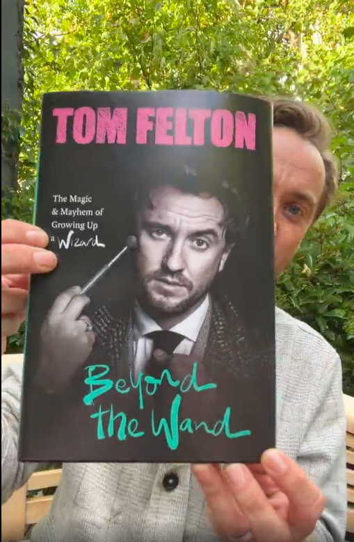 Tom Felton holds up the front cover of his memoir, "Beyond the Wand." It features a black and white photo of Tom and the book title in cursive writing.