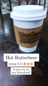 Sitting on a wood table, a white cup with a brown butterbeer cup cozy with Hot Butterbeer inside. The caption is Hot Butterbeer rating 5/5. A must try for any Potterhead.