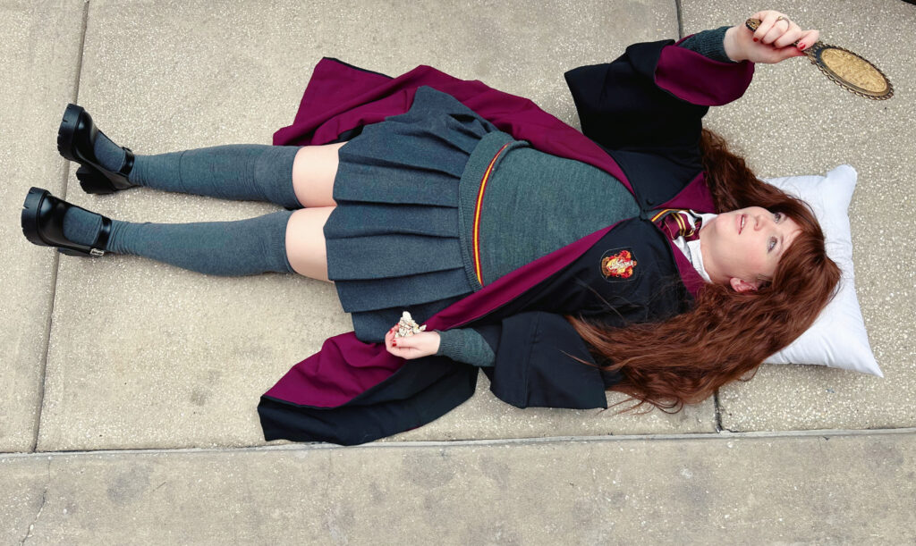 A Harry Potter fan lays on the ground, mirror in hand and head on a pillow, playing the part of petrified Hermione, after her basilisk attack