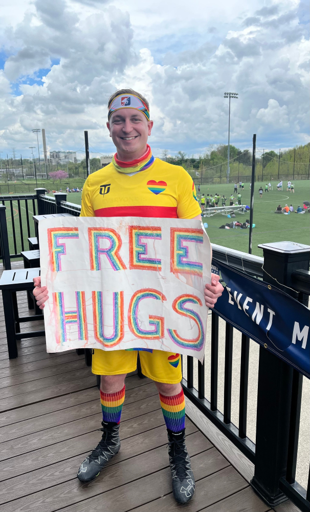 Richard is shown in a yellow and rainbow outfit, holding a sign with "free hugs" written on it in rainbow-colored capital letters.