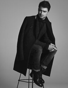 Black and white image of Daniel Radcliffe wearing a suit with a sweater and a bowtie, broodingly staring into the distance