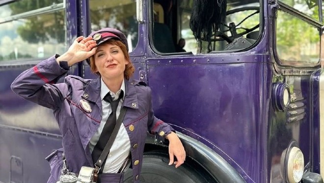 The Knight Bus Conductor tips her cap while leaning on the front wheel.