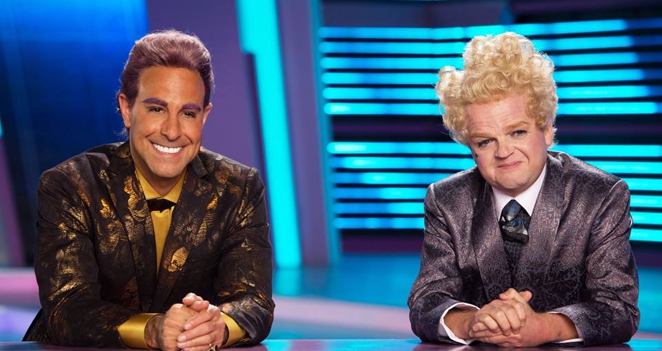 Stanley Tucci as Caesar Flickerman and Toby Jones as Claudius Templesmith sitting at a desk in "The Hunger Games: Catching Fire"